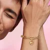 A close-up shot of a person's arm wearing a gold bracelet with a four-leaf clover charm, against a pink background. Produktbild-Vorschau