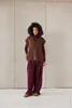 A woman stands against a concrete wall, wearing a brown knitted vest over a burgundy turtleneck and matching wide-leg pants. Produktbild-Vorschau