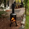 A black outdoor fireplace with flames is shown in a garden setting, with two people sitting at a table in the background. Produktbild-Vorschau