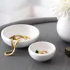 Two white ceramic bowls on a gray surface, one containing gold jewelry and the other a ring with a green stone. A white vase with a white flower is in the background. Náhľad obrázku produktu