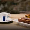 A white Lavazza coffee cup and saucer sit on a wooden table next to slices of cake on a white plate. Produktbild-Vorschau