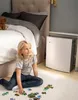 A young girl is sitting on the floor in a bedroom, playing with a puzzle, with an air purifier next to her and a bed in the background. Produktbild-Vorschau