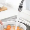 A faucet extender is shown attached to a kitchen faucet, with water running into a colander filled with small orange fruits in a sink. Produktbild-Vorschau