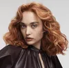 A woman with red wavy hair is shown from the chest up, wearing a dark leather top. Anteprima dell’immagine prodotto