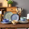 A collection of decorative ceramic plates and bowls with intricate patterns, arranged on a wooden table. The background includes a wooden cabinet, a potted plant, and dried flowers. Produktbild-Vorschau