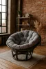 A papasan chair with a gray cushion sits on a rug in a room with exposed brick walls and a large window. Produktbild-Vorschau