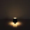 A small, black table lamp with a beige base is lit, casting a warm glow on a flat surface. Produktbild-Vorschau