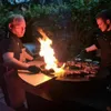 Two men are grilling steaks over a large, circular fire pit with flames erupting. Produktbild-Vorschau