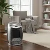 A ceramic heater is placed on a wooden floor in a living room, next to a chair and a plant. Podgląd zdjęcia produktu