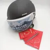 A black ski helmet with a printed face mask attached, and several red instruction booklets are placed in front of it on a white surface. Náhled obrázku produktu