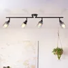 A black track light fixture with four adjustable spotlights is mounted on a white ceiling above a textured abstract painting and a hanging potted plant. Produktbild-Vorschau