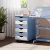 A blue and white roll container with drawers next to a blue desk with drawings on it, in a room with a window and a plant. Produktbild-Vorschau