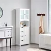 A white bathroom cabinet with glass doors and drawers is shown in a bathroom setting. Podgląd zdjęcia produktu