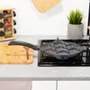 A frying pan with seven circular indentations is placed on a gas stove burner. The pan has a speckled black and gray granite coating and a black handle. To the left of the pan, a wooden cutting board and a spice rack are visible. A potted green plant sits  Anteprima dell’immagine prodotto