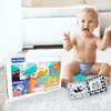 A happy baby is sitting on a white rug next to a box of baby cloth books and a separate open cloth book. Anteprima dell’immagine prodotto