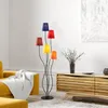 A colorful floor lamp stands in a modern living room next to a yellow sofa and a wooden cabinet. Produktbild-Vorschau