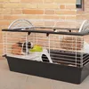A guinea pig cage with a guinea pig inside, placed on a tiled surface in front of a brick wall. Podgląd zdjęcia produktu