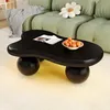 A black coffee table with a unique cloud shape and spherical legs sits on a cream rug in a living room. The table has a magazine and a bowl of tangerines on it. A green sofa with a flower-shaped pillow is visible in the background. Voir les images du produit