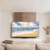 A modern living room with a large TV displaying a beach scene, mounted above a white media console. A decorative wooden panel is behind the TV. Produktbild-Vorschau