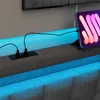 A close-up view of a bed frame with integrated USB charging ports and LED lighting, featuring a tablet plugged into one of the ports. Produktbild-Vorschau