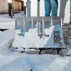 A snow shovel filled with snow, resting on a wooden deck with a person in jeans in the background. Produktbild-Vorschau
