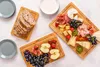 A top-down view of three bamboo cutting boards filled with various food items, including bread, cheese, meats, fruits, and nuts, arranged on a light-colored surface. Produktbild-Vorschau