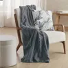 A dark gray blanket and a floral pillow are draped over a white armchair, with a round ottoman to the left and a wooden bench with books in the background. Produktbild-Vorschau