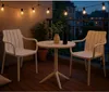 A small round table and two chairs on a balcony at dusk, with string lights overhead. Náhľad obrázku produktu