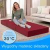 A woman sits on a red foldable mattress in a room with a couch and curtains. Anteprima dell’immagine prodotto