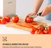 A person is slicing tomatoes on a bamboo cutting board, with several whole tomatoes in the foreground and a box with a logo in the background. The bottom part of the image contains text and an icon. Produktbild-Vorschau
