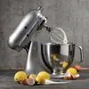 A silver KitchenAid stand mixer with a whisk attachment is shown in use, with lemons and eggshells scattered around the base. Podgląd zdjęcia produktu
