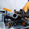 A person wearing a tan glove is using a reciprocating saw to cut a gray pipe. The saw has black and orange accents, and the brand name "WORX" is visible on the equipment in the background. Voir les images du produit
