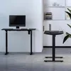 Two black height-adjustable desks are shown in a modern office setting with a white wall and light gray floor. One desk has a monitor, keyboard, and mouse, while the other has a laptop and mouse. Náhled obrázku produktu