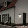A black outdoor wall light is mounted on a white brick house next to a black door. Produktbild-Vorschau