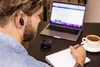 A man wearing earbuds is working on a laptop, taking notes in a notebook, and drinking coffee. Produktbild-Vorschau