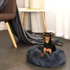 A small dog is resting in a plush, round dog bed next to a chair and a small table. Anteprima dell’immagine prodotto