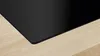 A close-up view of the corner of a black induction cooktop resting on a light wood surface. Produktbild-Vorschau