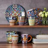 Auf einem Holzregal vor grauer Wand stehen bunte Geschirrteile und eine Vase mit weißen Blumen. Náhled obrázku produktu