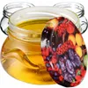 A glass jar filled with honey, with a decorative lid featuring a pattern of various fruits. Several empty glass jars are visible in the background. Produktbild-Vorschau