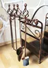 A fireplace tool set and log holder are displayed against a stone wall and wooden floor. Podgląd zdjęcia produktu