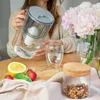 A person is pouring water from a filter pitcher into a glass, with fruit and flowers on a table. Náhled obrázku produktu