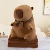 A plush capybara toy sitting on a folded blanket, with small decorative trees and a rainbow in the background. Podgląd zdjęcia produktu