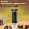 A portable coffee machine is displayed on a wooden table with coffee beans and capsules around it. The background is blurred and appears to be outdoors. Podgląd zdjęcia produktu
