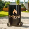 A black metal fire pit with a gold flame logo and text 'Grillrost - BBQ' is shown outdoors on a paved patio, with a garden and trees in the background. Produktbild-Vorschau