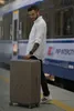 A man stands on a train platform holding a suitcase next to a train. Produktbild-Vorschau