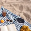 A portable CD player, headphones, a book, sunglasses, a drink, and a sweater are laid out on a striped blanket on a sandy beach. Podgląd zdjęcia produktu