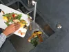 A person is scraping food scraps from a cutting board into a kitchen sink with a garbage disposal. Produktbild-Vorschau
