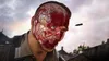 A close-up of a person's face with exposed muscles and blood, a bullet flying past, and a village in the background. Podgląd zdjęcia produktu