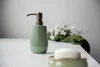A green ceramic soap dispenser with a bronze pump sits on a white surface, with a blurred green cloth and white flowers in the background. Produktbild-Vorschau