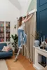 A woman is standing on a ladder and adjusting curtains in a living room. Náhľad obrázku produktu